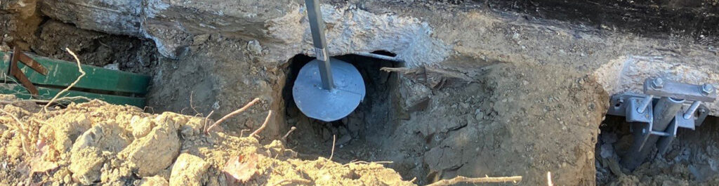 Close-up of a helical pier being driven into the soil beside a brick foundation wall during foundation repair and stabilization work.