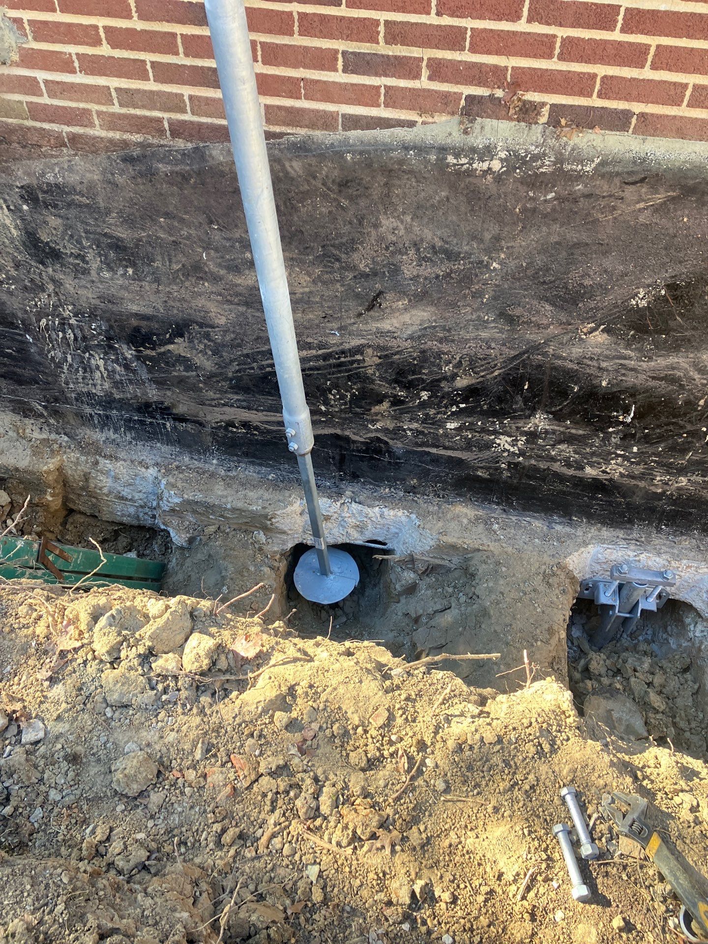 Close-up of a helical pier being driven into the soil beside a brick foundation wall during foundation repair and stabilization work.